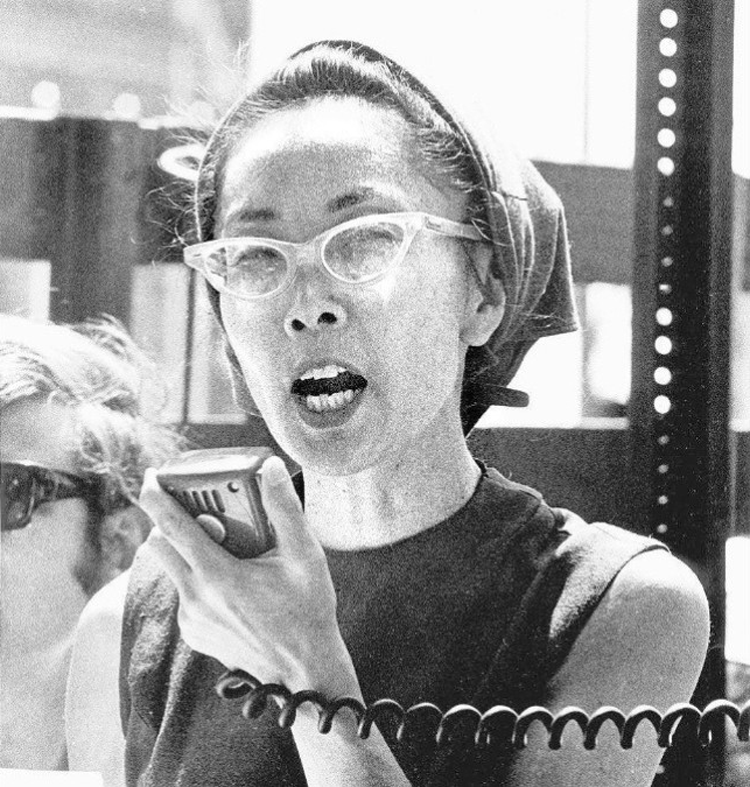 Photo Description: the most common pic of Yuri Kochiyama where she is speaking into a microphone, while wearing tear drop shaped glasses, and a rag over her head.